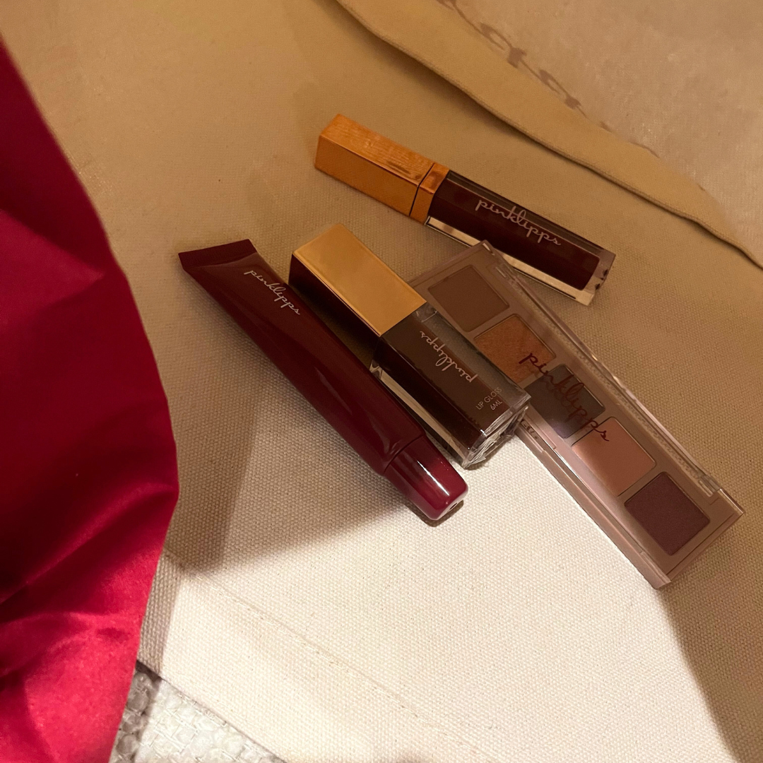 Pink Lipps Tote Bag lifestyle flat-lay with burgundy lipstick, lip balm and eyeshadow — tan neutral bag with burgundy straps styled on light background